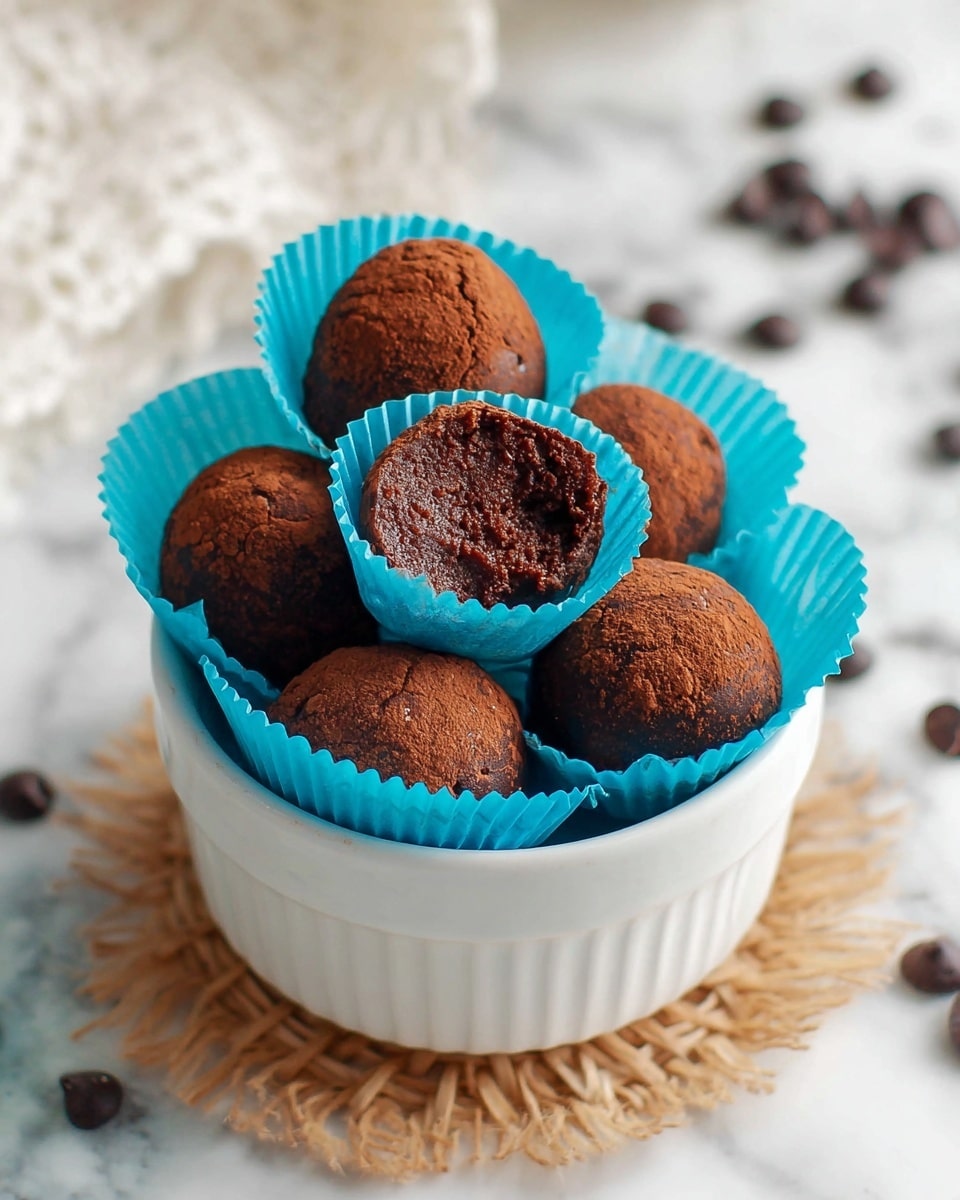 A white cup filled with five dark brown chocolate balls placed in blue paper cups, showing a textured cracked surface; one ball is cut in half to reveal its rich, slightly crumbly inside, all sitting on a light-colored woven coaster on a white marbled surface, with a few chocolate pieces scattered nearby, soft light highlighting the smooth and cracked textures, photo taken with an iphone --ar 4:5 --v 7