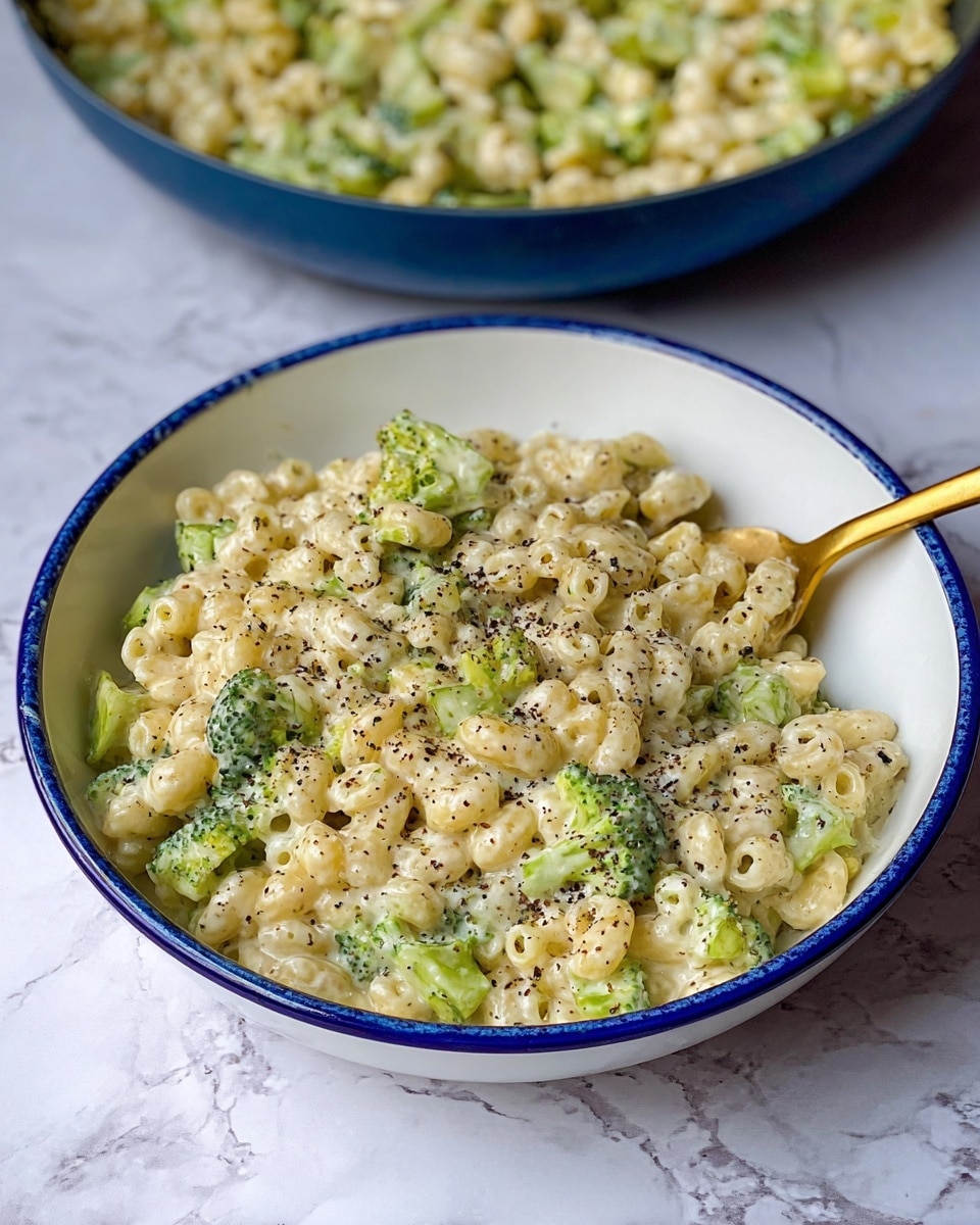 A white bowl filled with creamy pasta mixed with small pieces of green broccoli, the pasta is tubular and light beige, covered in a smooth white sauce with black pepper specks sprinkled on top. A gold spoon holds a scoop of the pasta and broccoli mixture above the bowl, the spoon handle extends to the right. In the blurred background, there is a larger white bowl filled with the same dish sitting on a white marbled surface. photo taken with an iphone --ar 4:5 --v 7