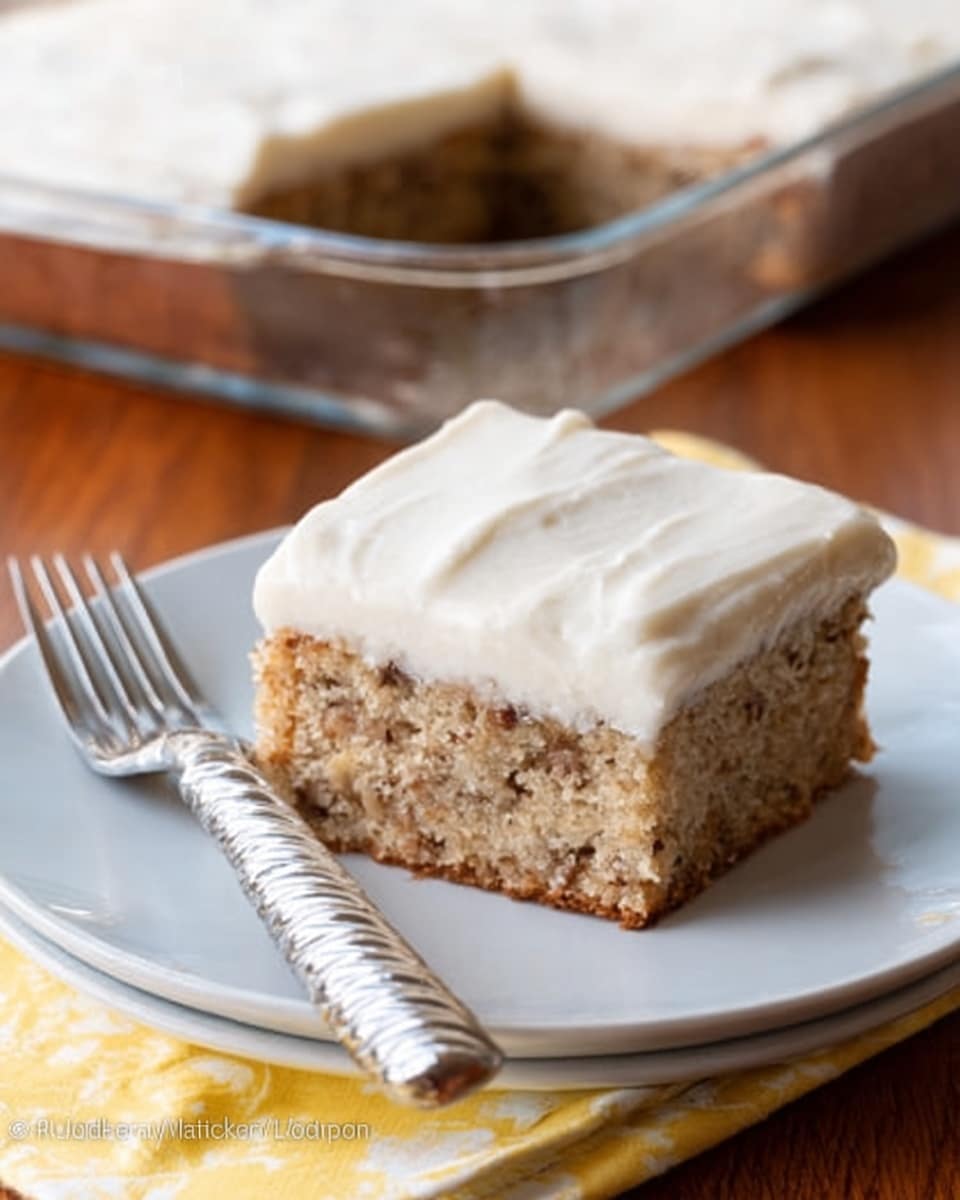 A single square piece of light brown cake with tiny bits visible inside sits centered on a plain white plate. The cake is topped with a thick, smooth, creamy white layer of frosting that covers it evenly. Next to the cake on the plate is a silver fork with a swirled handle resting diagonally. The plate is on a wooden surface, and the background shows a larger cake in a clear glass dish. The overall look is simple and inviting, with natural colors and soft lighting. Photo taken with an iphone --ar 4:5 --v 7