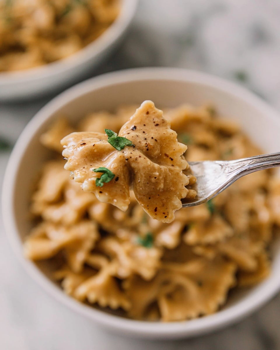 A close-up view of creamy pasta in a white bowl on a white marbled surface, with a silver fork holding a bite of pasta in the foreground. The pasta has wavy edges and is coated in a light brown sauce with small green herb leaves and specks of black pepper on top, giving it a rich and smooth texture. The background bowl is full of the same saucy pasta, softly blurred to focus on the pasta on the fork. photo taken with an iphone --ar 4:5 --v 7