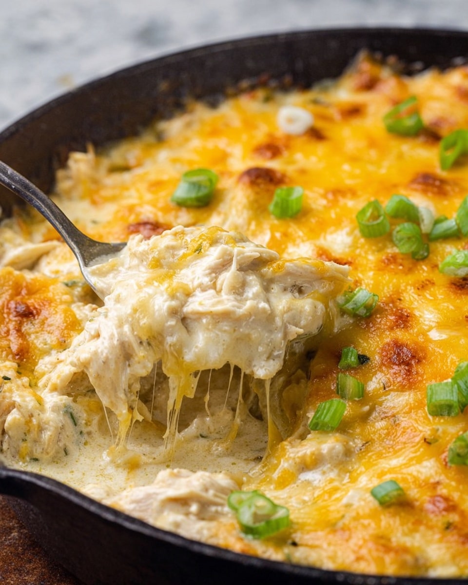 This close-up view shows a round black skillet filled with a creamy dish featuring several layers. The bottom layer is a smooth white sauce mixed with shredded light brown chicken, covered by a melted cheese layer that is golden yellow and slightly browned in spots. Small green onion pieces are scattered over the cheese, adding bright green pops of color. The cheese looks stretchy and gooey, blending with the creamy sauce below. The skillet has a metal spoon lifting some of the cheesy chicken mixture, with a white marbled texture in the background. photo taken with an iphone --ar 4:5 --v 7
